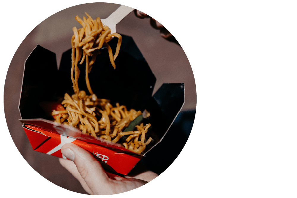 Two hands holding a cardboard takeaway box filled with noodles and a white plastic fork.