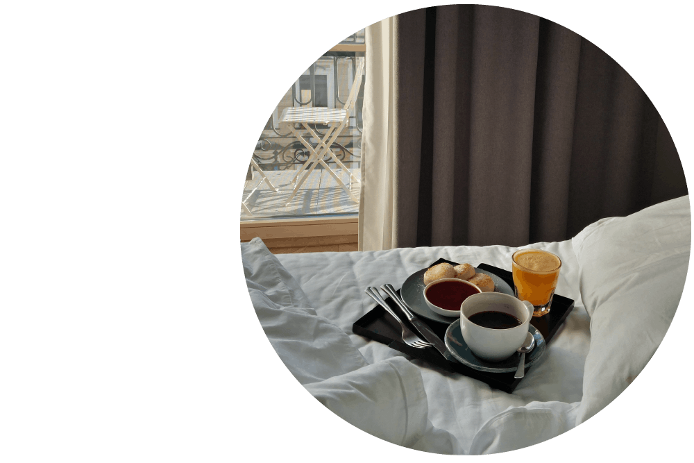 A tray with coffees, pastry and cutlery resting on an unmade hotel bed with white sheets and a window with a balcony in the background.