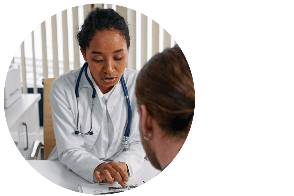 A doctor wearing a white coat and stethoscope sitting behind a desk. They are looking down at a document, pointing to the page and speaking with a thoughtful expression to a second person sitting across from them.