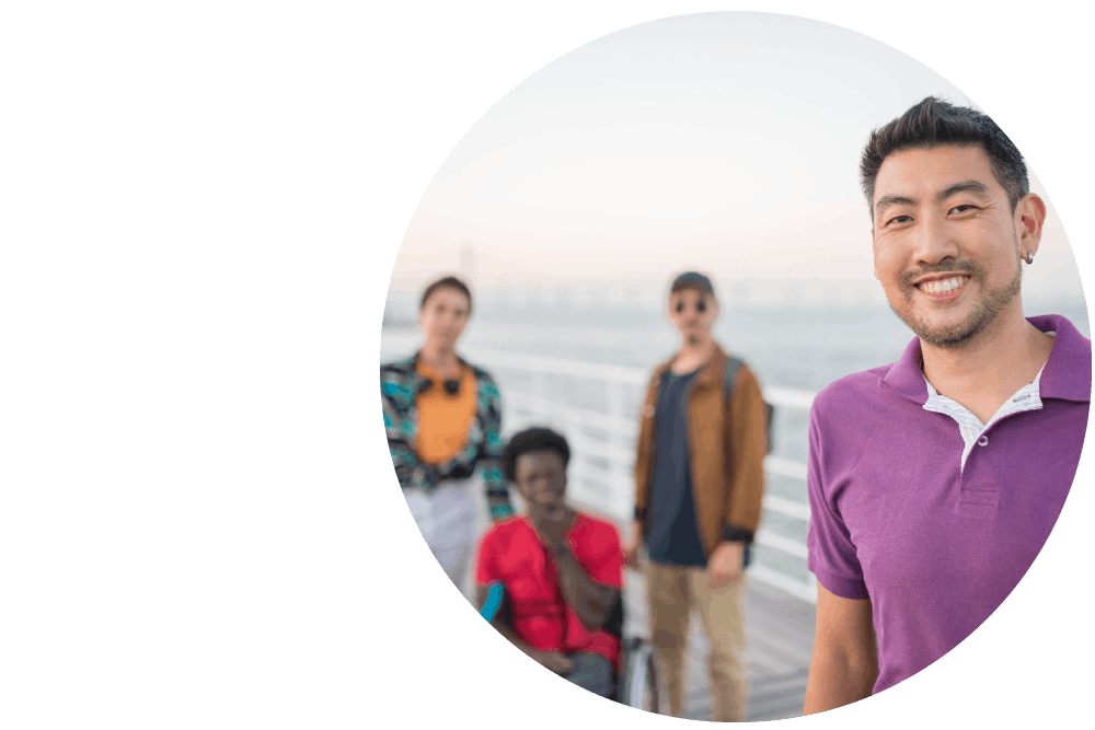 A diverse group of friends standing at a waterfront. One person in the foreground looks directly into the camera, smiling widely.