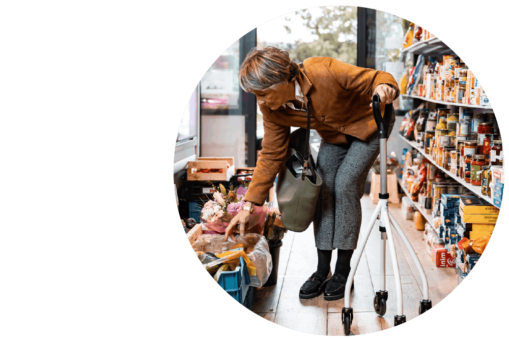 A person using a mobility aid, standing in the aisle of a supermarket. They are leaning down to retrieve products from a display with shelves in the background.