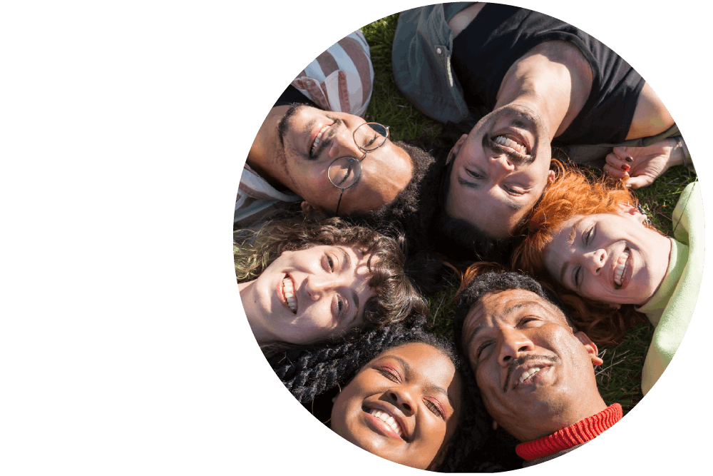 A diverse group of six smiling people, lying on the grass in the sunlight with their heads together forming a circle.