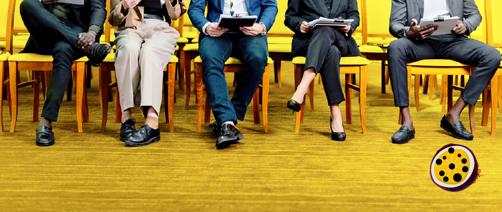A row of five people from the waist down, sitting in yellow chairs against a yellow background. They are dressed in business attire, some holding clipboards or papers, as if waiting to be interviewed. Passion4Social (logo).