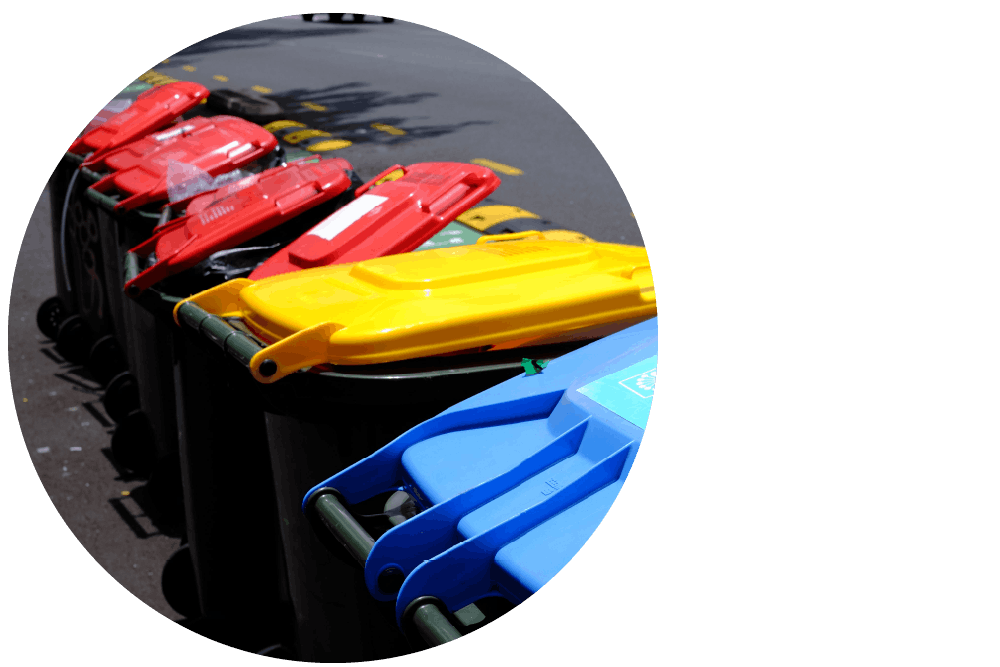 A row of outdoor recycling bins lined up on an asphalt street, with colourful blue, yellow, and red lids.