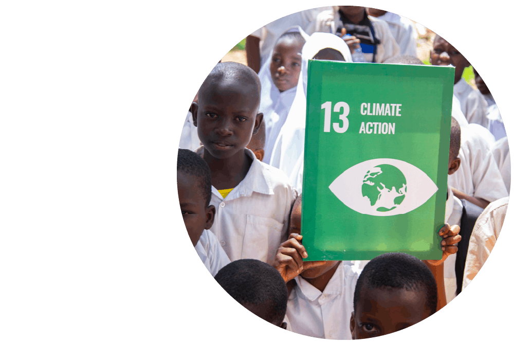 A group of schoolchildren standing together outdoors, with one child holding a green placard for Sustainable Development Goal 13. The placard reads ‘13: Climate Action’, with a white graphic of a globe inside an eye.