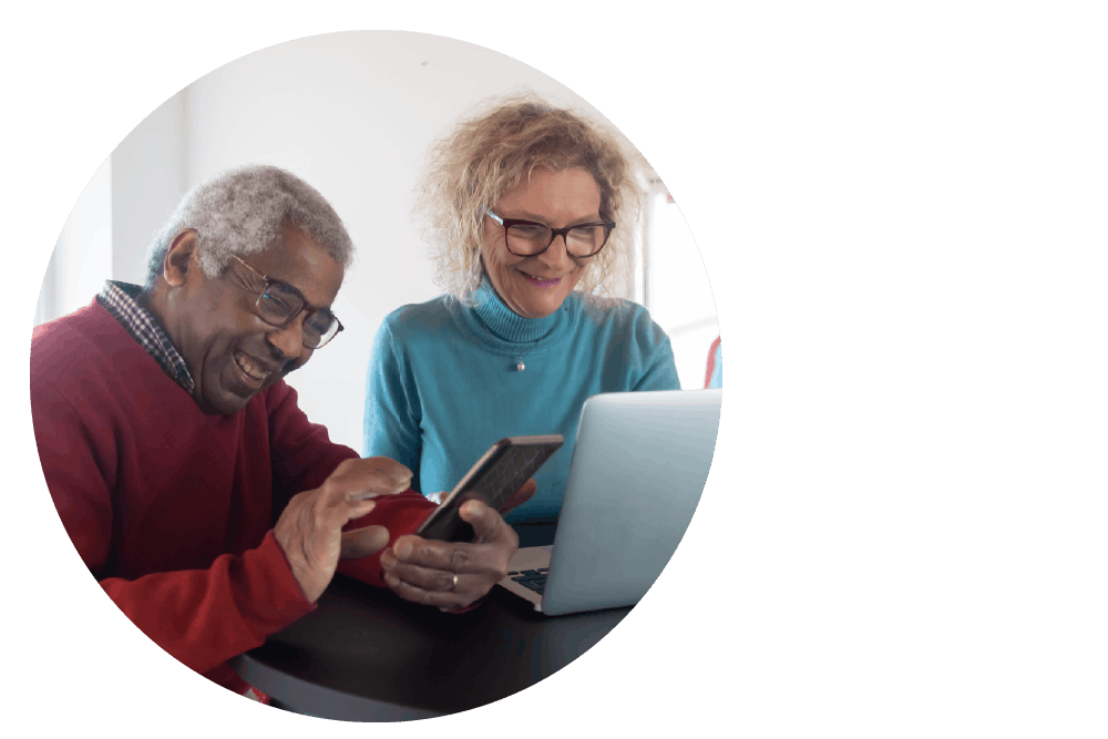 An older couple smiling together while using a smartphone and laptop.